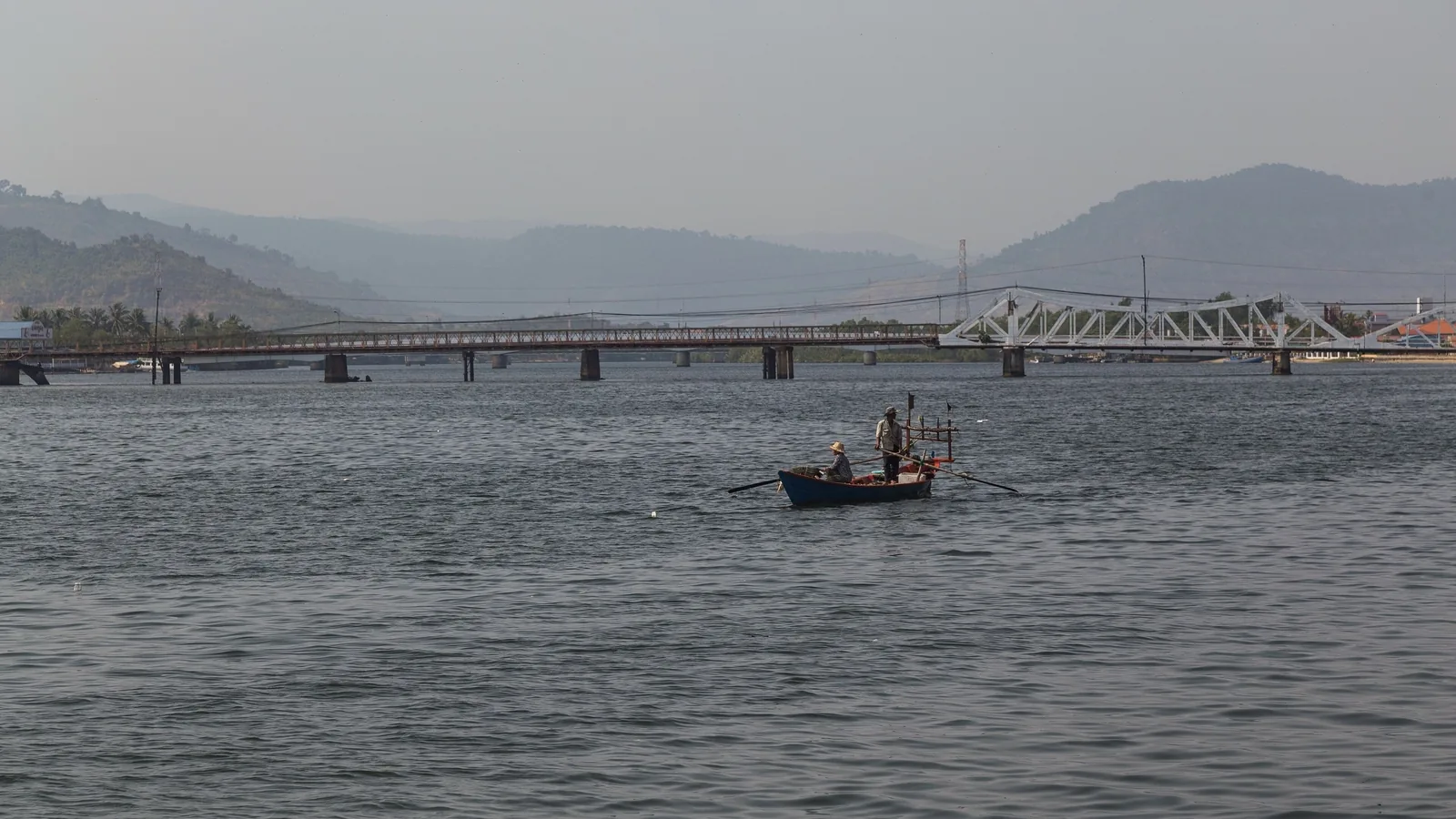 A small boat with two fishermen crosses the Praek Tuek Chhu river near Kampot's bridge and misty hills.