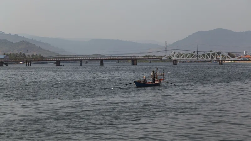A small boat with two fishermen crosses the Praek Tuek Chhu river near Kampot's bridge and misty hills.