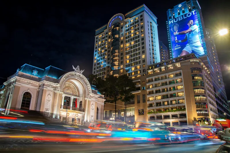 Long-exposure night traffic streaks past the illuminated Saigon Opera House and skyscrapers in Ho Chi Minh City.