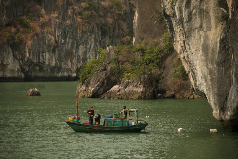 Fishermen work on a small wooden boat between towering limestone karsts in Bai Tu Long Bay.