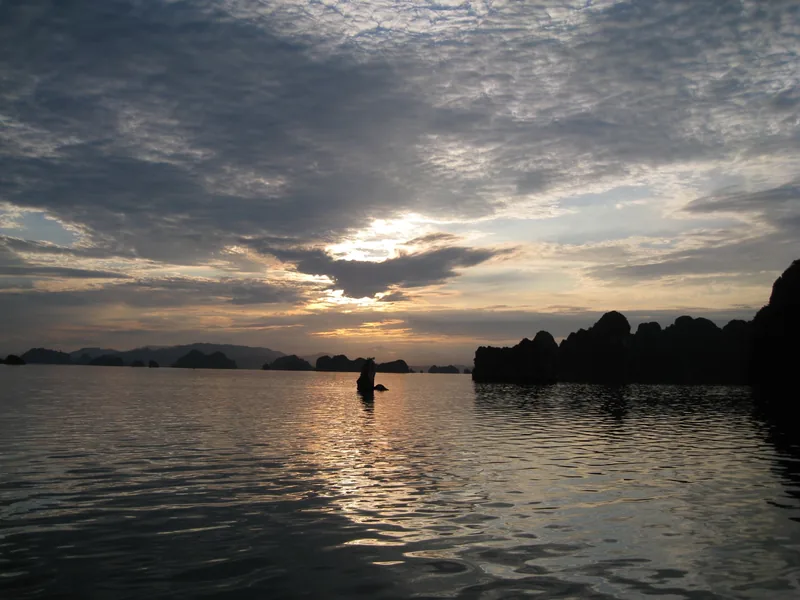 Sun sets behind silhouetted limestone karsts over calm waters of Bai Tu Long Bay.