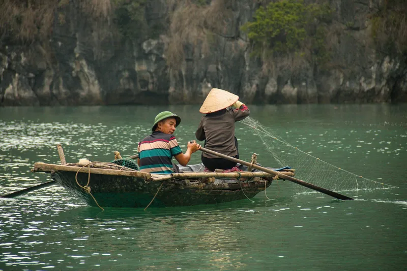 Two fishers cast a net from a small wooden boat on the emerald waters of Bai Tu Long Bay.
