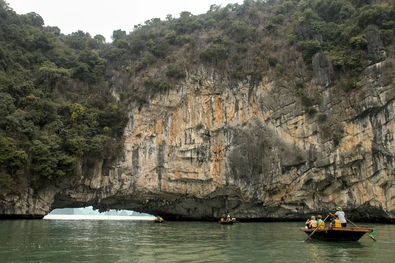Small rowing boats pass beneath a towering limestone cliff with a sea arch in Bai Tu Long Bay