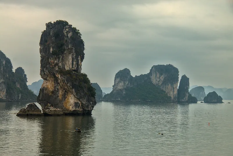 Limestone karsts rise from calm waters under a misty sky in Bai Tu Long Bay, with a small boat below.