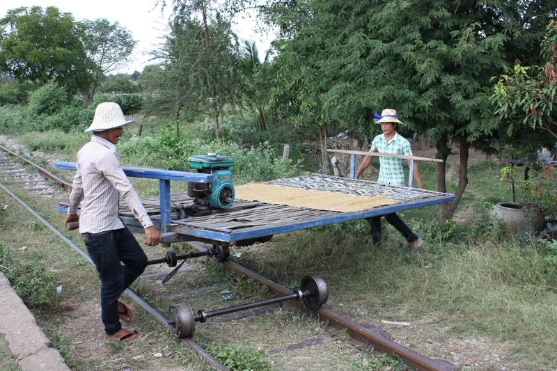 Two men push a bamboo-platform bamboo train cart onto rails amid greenery in Battambang, Cambodia.
