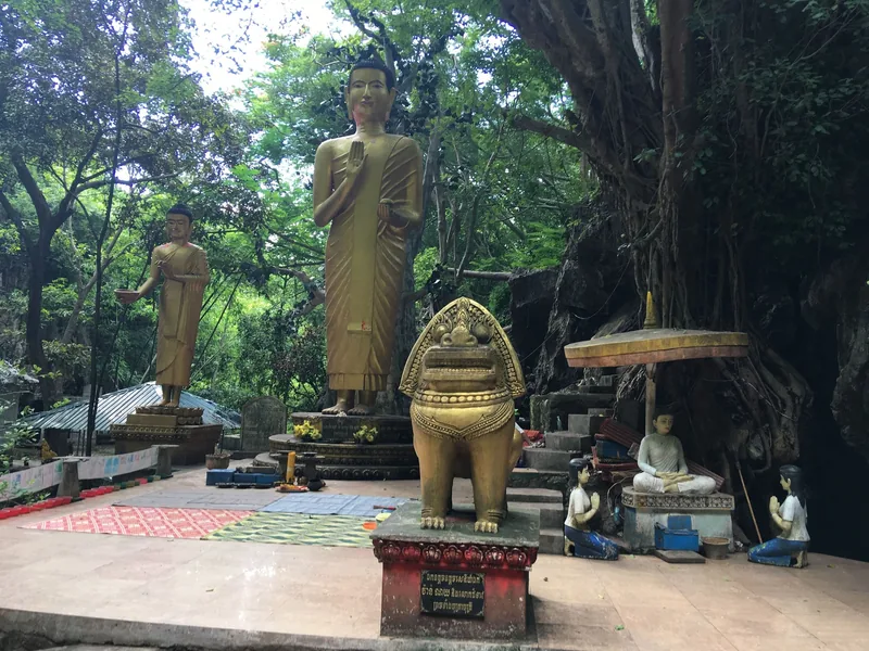Golden Buddha statues and a lion figure stand among trees on Phnom Sampeau near Battambang.