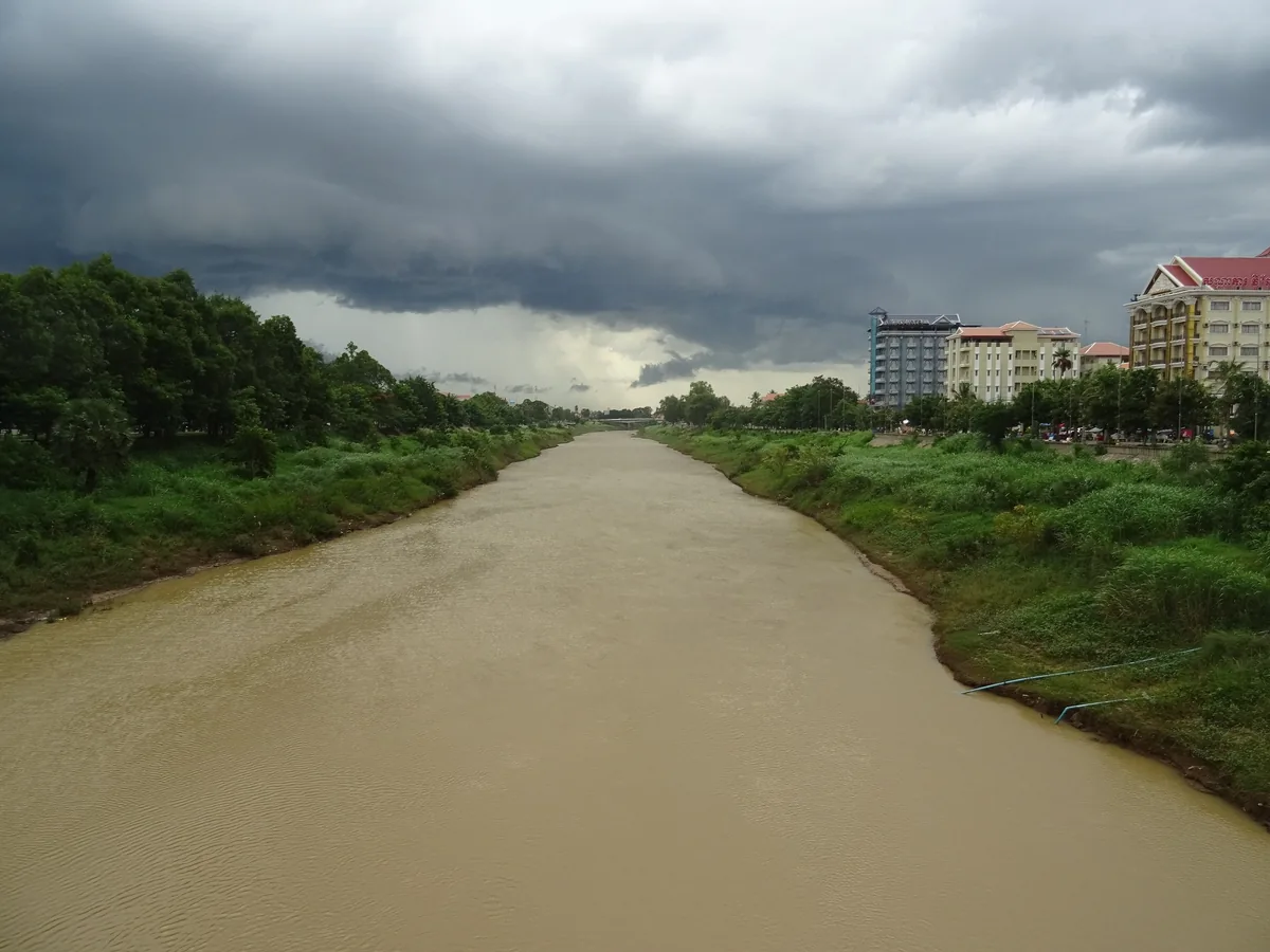 Storm clouds gather over the muddy Sangker River flowing through Battambang, lined with trees and low buildings.