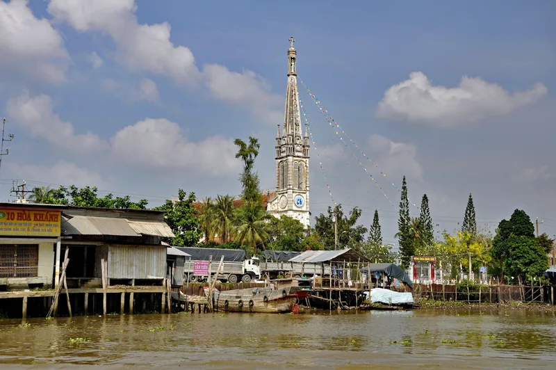 A tall Gothic church spire rises above riverside stilt houses along a Mekong Delta waterway near Cai Be.