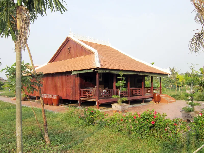 A traditional wooden stilt house with a red tiled roof stands in a garden in the Mekong Delta.