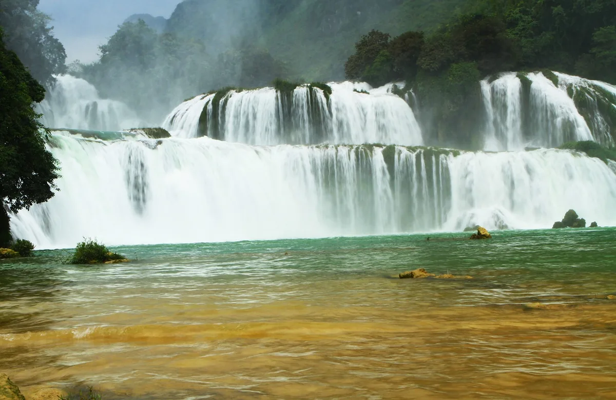 Wide tiered curtains of white water cascade over limestone cliffs into a turquoise pool at Ban Gioc Waterfall.