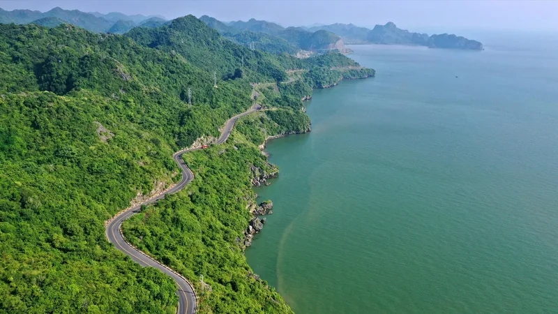 A winding coastal road cuts through forested hills above the turquoise waters of Lan Ha Bay.
