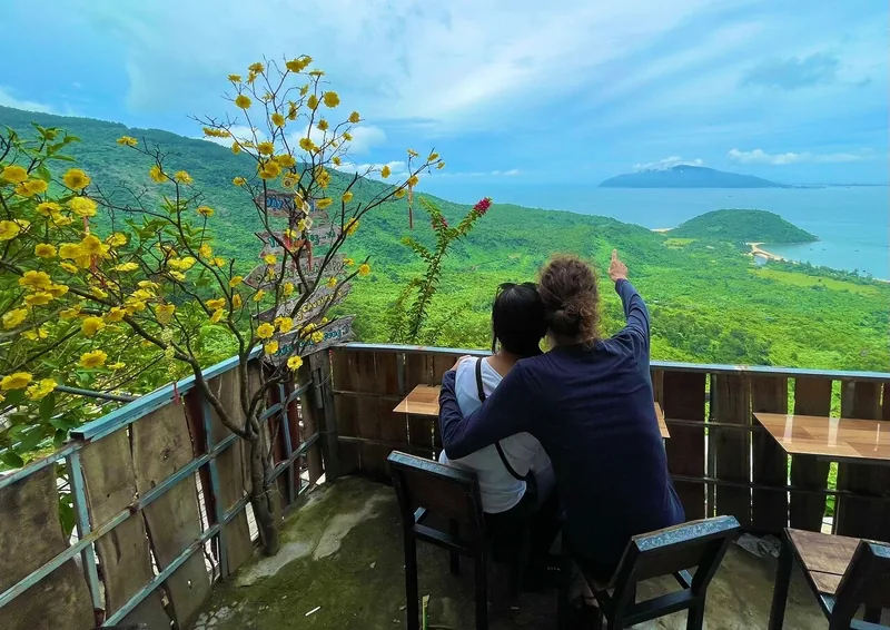 A couple sits at a hillside viewpoint café overlooking green hills and the coast along the Hue to Hoi An road.