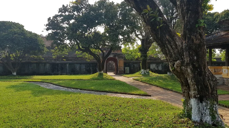 A shaded lawn with old trees leads to an arched gate in the ancient walls of the Hue Imperial City.