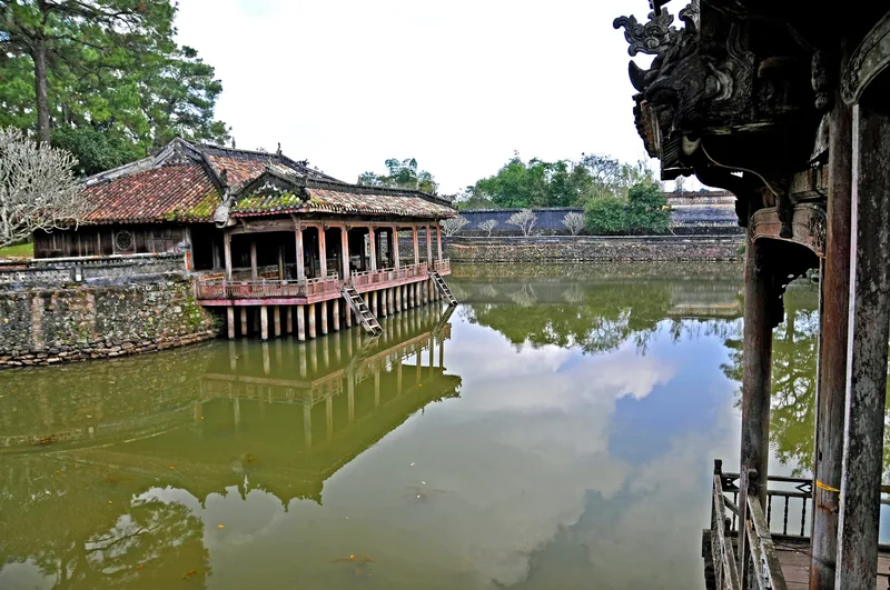 A traditional wooden pavilion on stilts reflects in a calm lake within a historic Hue royal tomb complex.