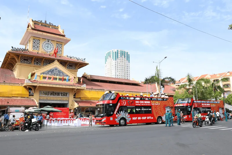 Red hop-on hop-off double-decker buses parked in front of the yellow clock tower of Binh Tay Market in Ho Chi Minh City.