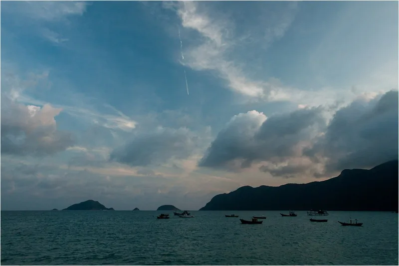 Small fishing boats float on calm water beneath a dramatic cloudy sky near Con Dao's coastal mountains.
