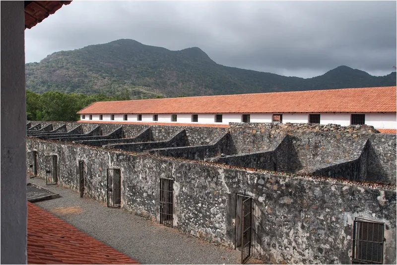 Rows of stone prison cells with iron-barred doors line the old French colonial prison at Con Dao, mountains behind.