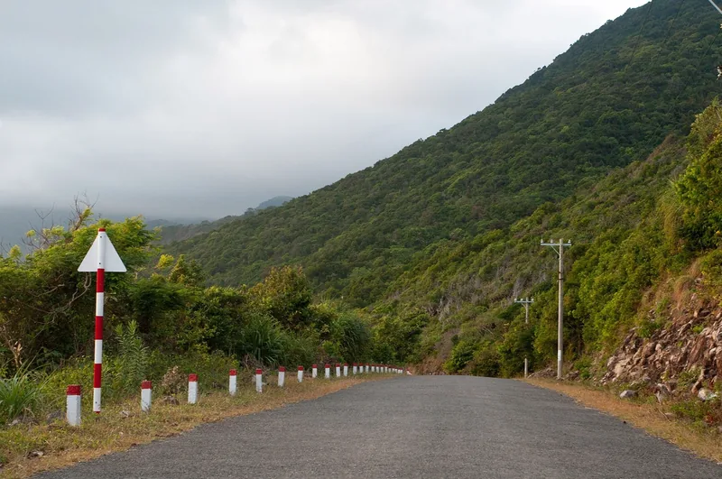 A quiet mountain road with red-and-white guardrail markers winds through forested hills in Con Dao.