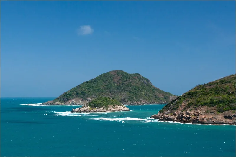A green forested islet rises from turquoise waters near Con Dao, with waves breaking on rocky shores.