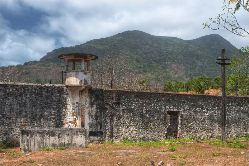 A weathered guard tower and stone wall stand at the historic prison site in Con Dao, mountains rising behind.