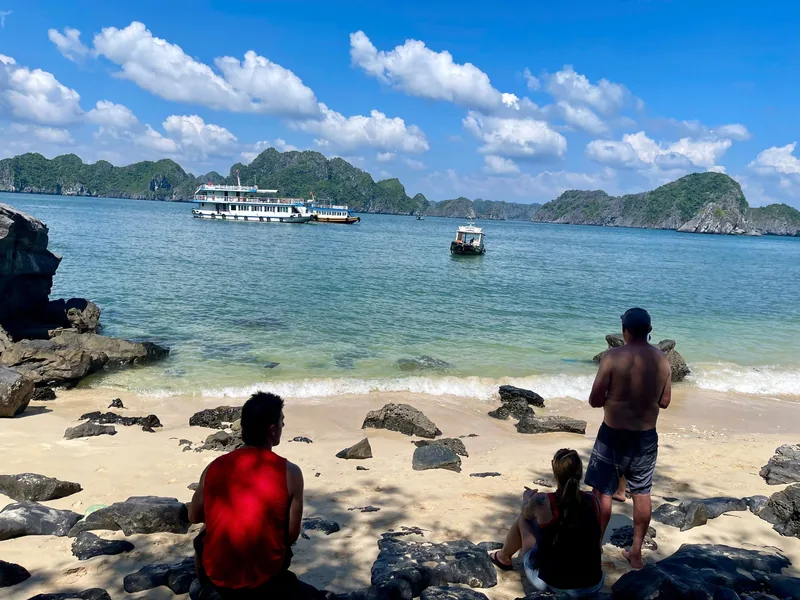 Visitors relax on a rocky beach watching cruise boats anchored among limestone islands in Lan Ha Bay.