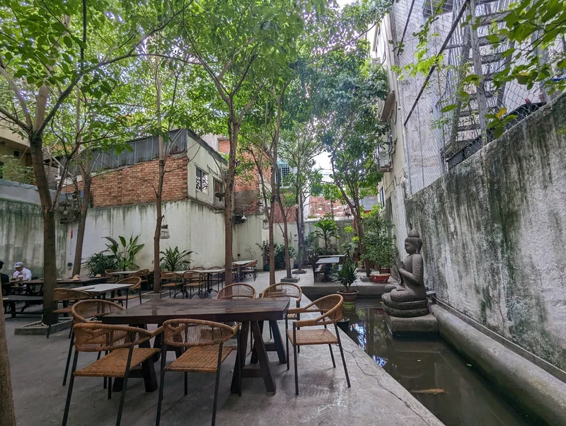 A shaded courtyard café with wooden tables, potted plants, and a small Buddha statue beside a narrow pond in Ho Chi Minh.