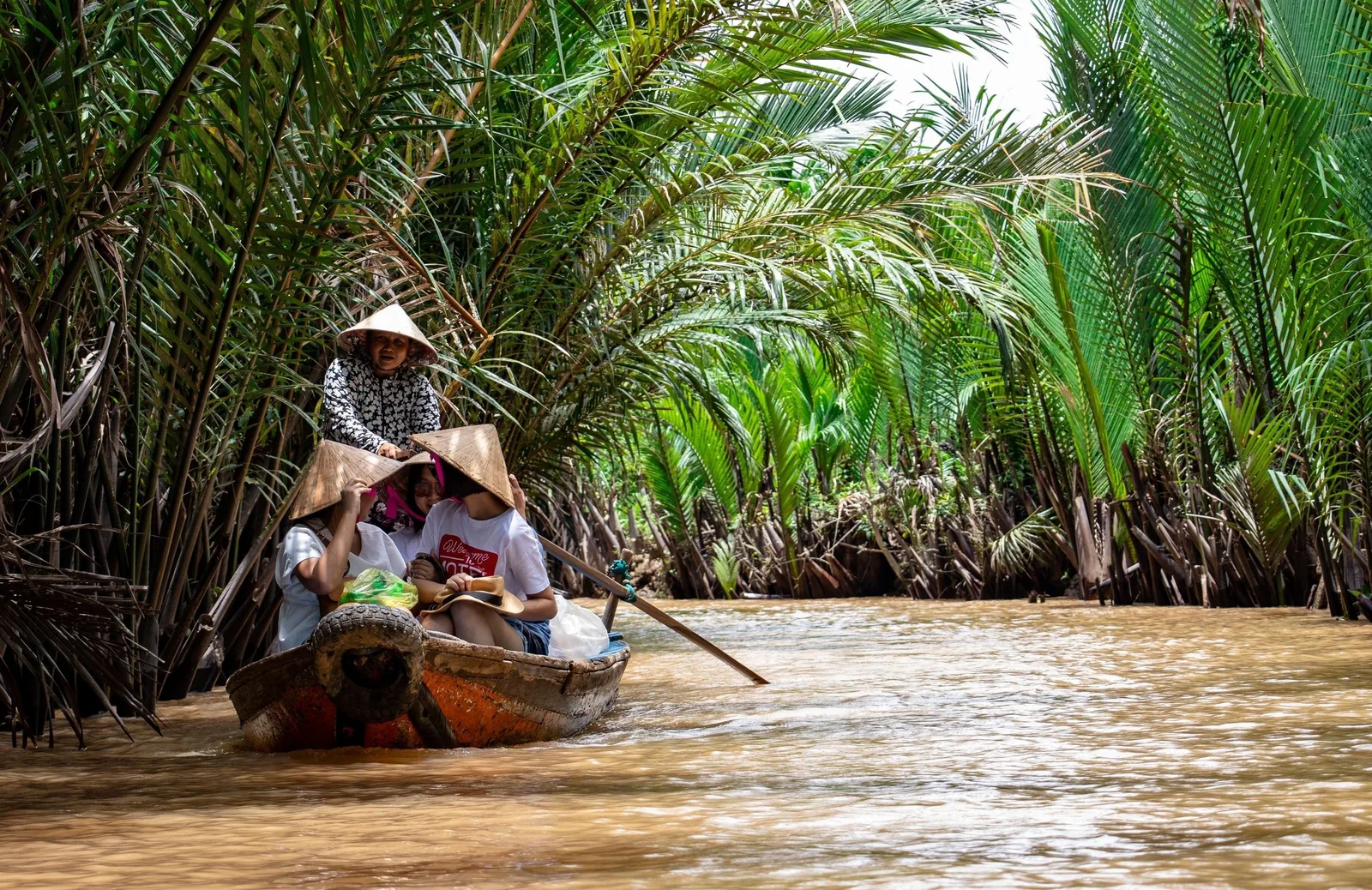 A rower guides tourists in conical hats through a narrow water palm channel on the Mekong Delta.