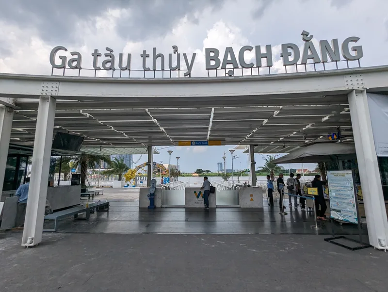 Entrance canopy of Bach Dang water bus station in Ho Chi Minh City with waiting passengers under a cloudy sky.