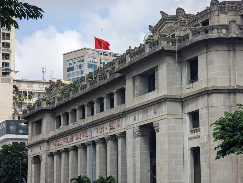 Ornate colonial stone facade of the State Bank of Vietnam building in Ho Chi Minh City with a red flag flying above.