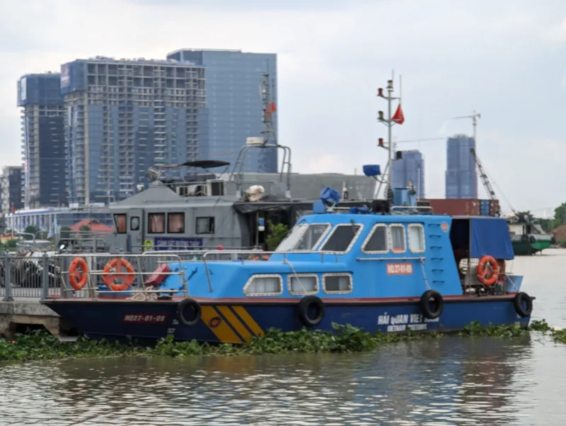 A blue Vietnam Customs patrol boat moored on the Saigon River with high-rise towers in the background.