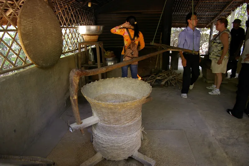 A traditional bamboo rice husking basket and mill sit under a thatched shelter as visitors look on near Cu Chi.