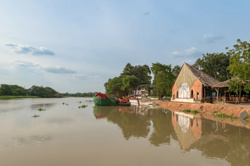 A wooden boat rests on the muddy bank of the Saigon River near Cu Chi, with a small riverside chapel among trees.