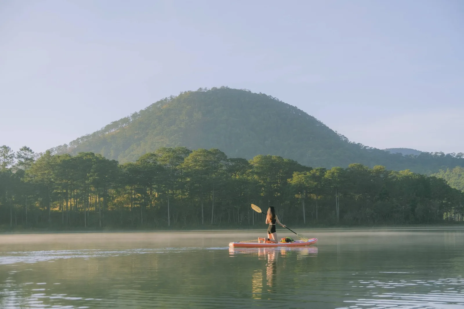 A paddleboarder crosses a misty lake beneath pine-covered hills near Da Lat.