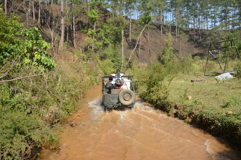 A jeep splashes through a muddy stream on a pine-forested hillside trail near Da Lat, Lam Dong.