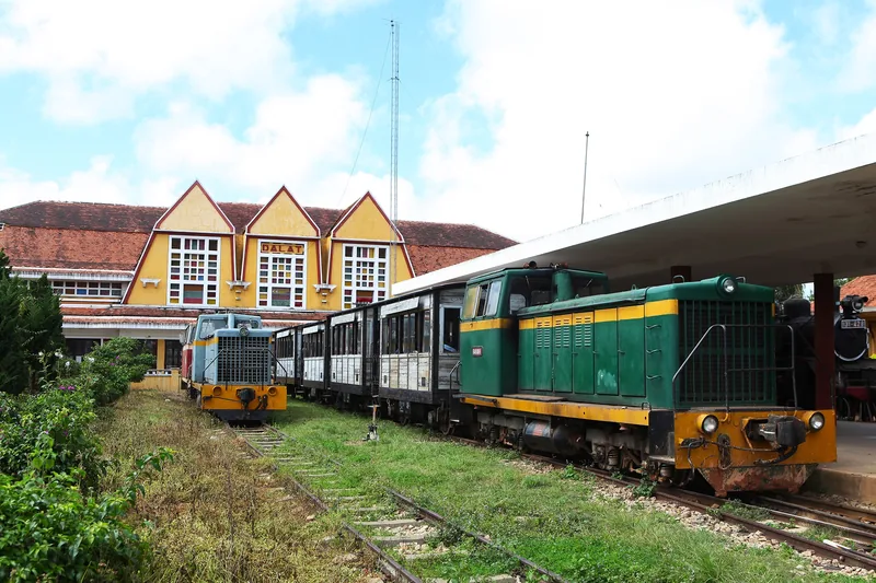 A green diesel locomotive sits on tracks beside the yellow triple-gabled Dalat railway station building.