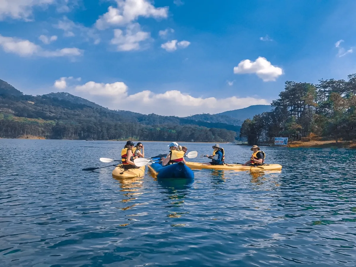 Kayakers in life jackets paddle yellow kayaks on Tuyen Lam Lake with pine-covered hills in Da Lat behind them.