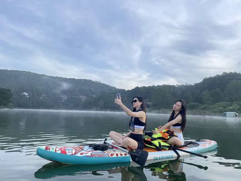 Two women sit on paddleboards taking a selfie on a calm misty lake surrounded by forested hills in Da Lat.