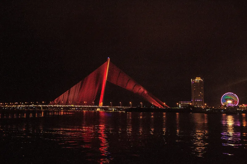 A red-lit cable-stayed bridge glows over the Han River in Da Nang at night, with a Ferris wheel nearby.