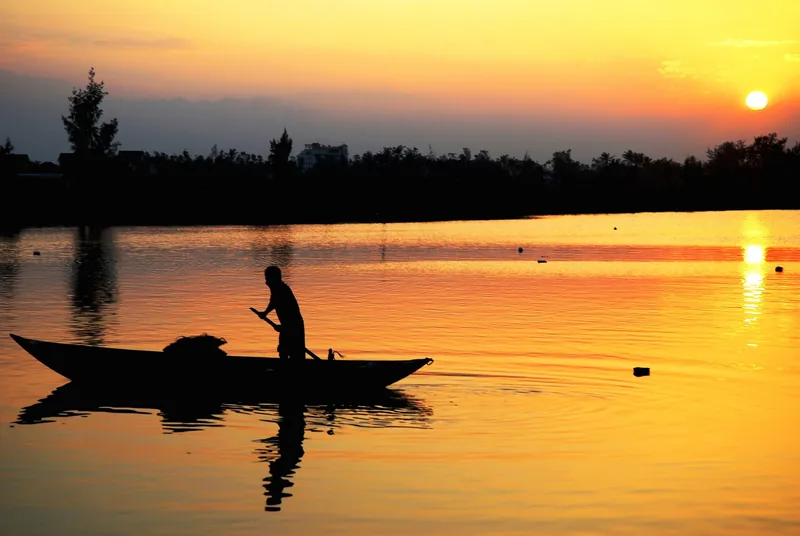 A boatman rows a small wooden boat across a calm river at golden sunset in Quang Tri.