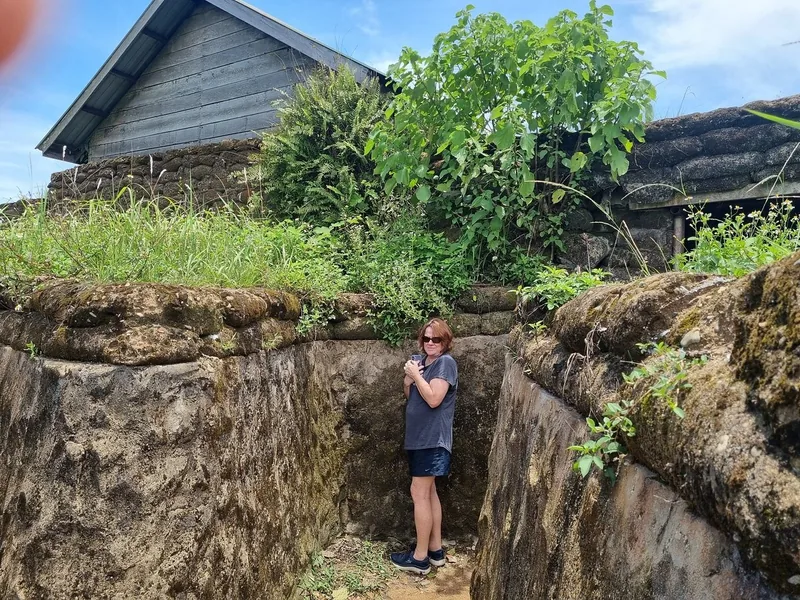 A visitor stands in an old earthen trench beside a wooden building on the Hue DMZ tour route.