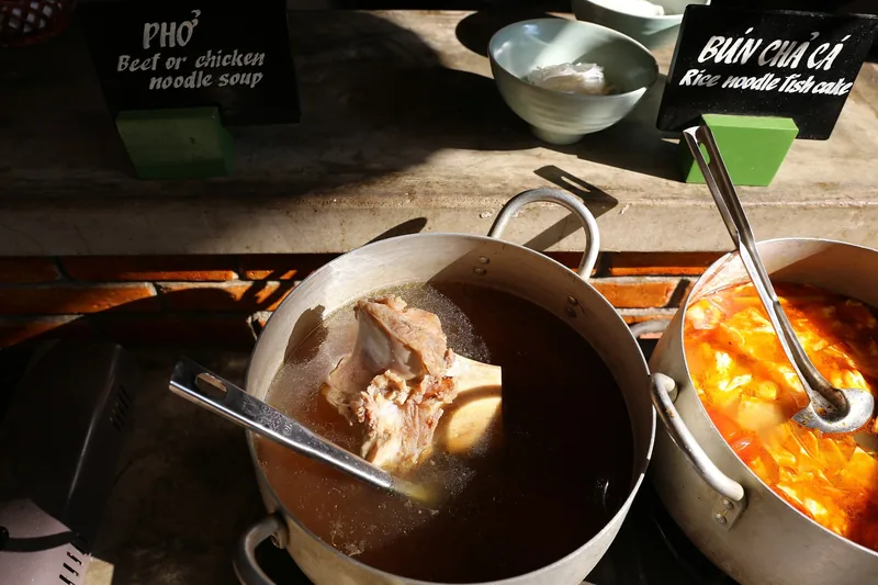 Pots of pho beef broth and bun cha ca fish noodle soup simmer at a Vietnamese food stall with menu signs.