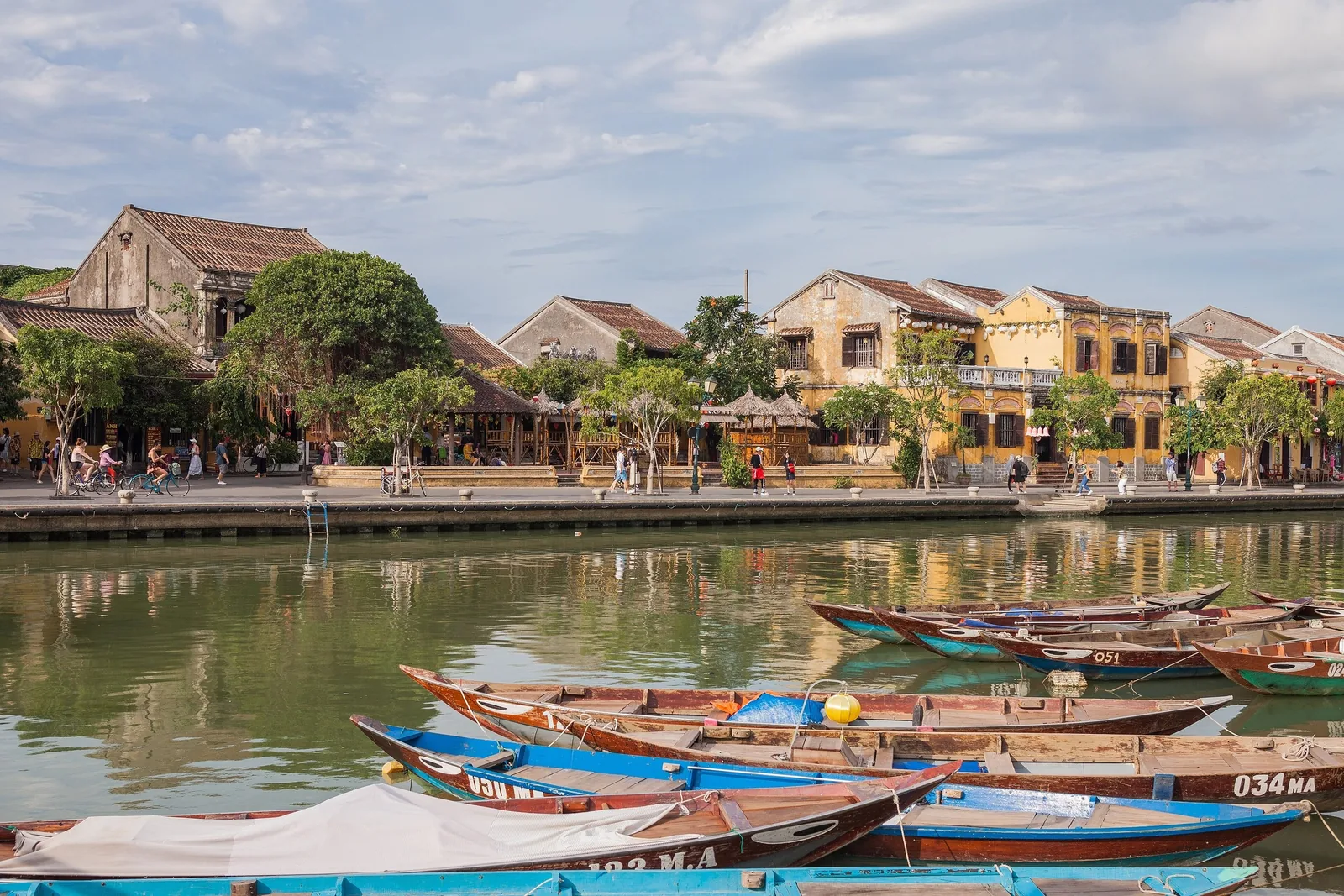 Wooden boats line the Thu Bon River in front of Hoi An's yellow colonial-era buildings and riverside promenade.