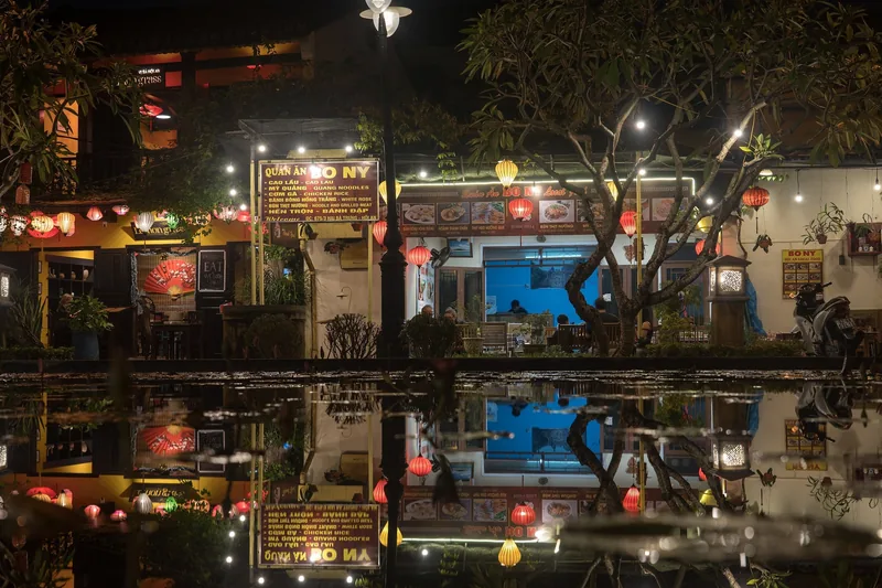 Colorful lanterns illuminate riverside restaurants in Hoi An's old town at night, their glow reflected in the water.