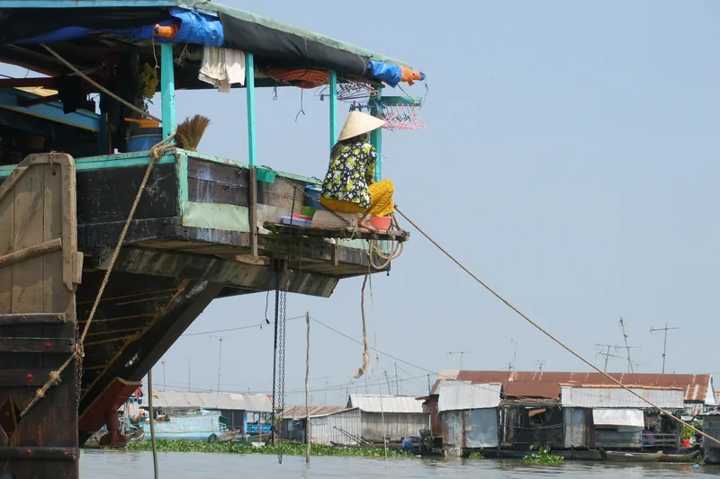 A woman in a conical hat sits at the back of a boat overlooking floating houses on the river in Chau Doc.