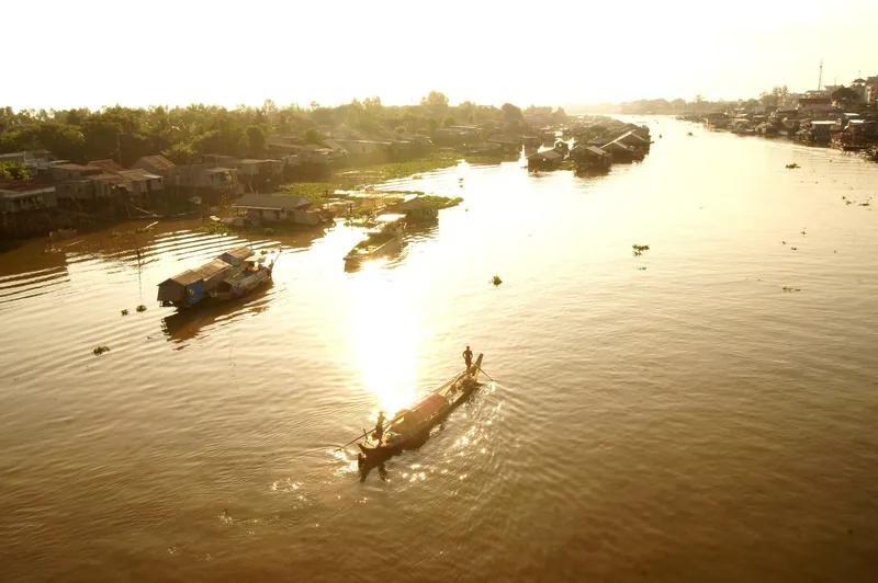 A rowboat glides across the golden Mekong River near riverside stilt houses at sunset in Chau Doc.