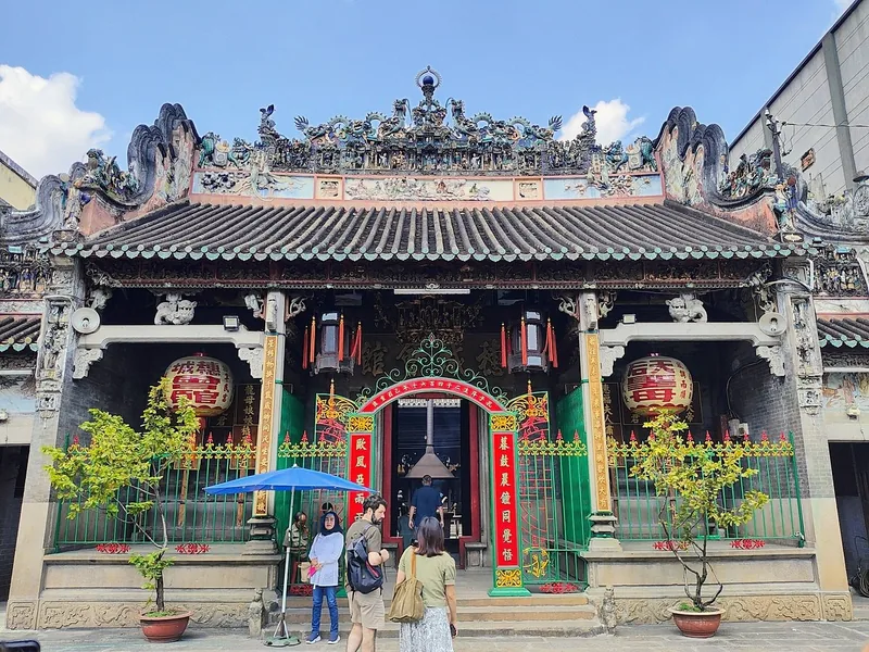 Ornate roof carvings and red lanterns adorn a Chinese-style temple facade in Saigon as visitors gather at the entrance.