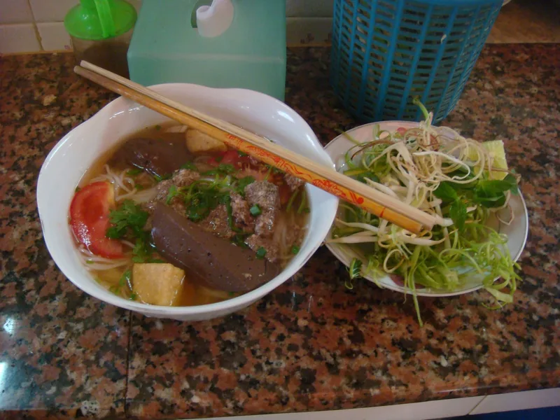 A bowl of bun rieu noodle soup with chopsticks and a side plate of fresh herbs on a table in Ho Chi Minh City.
