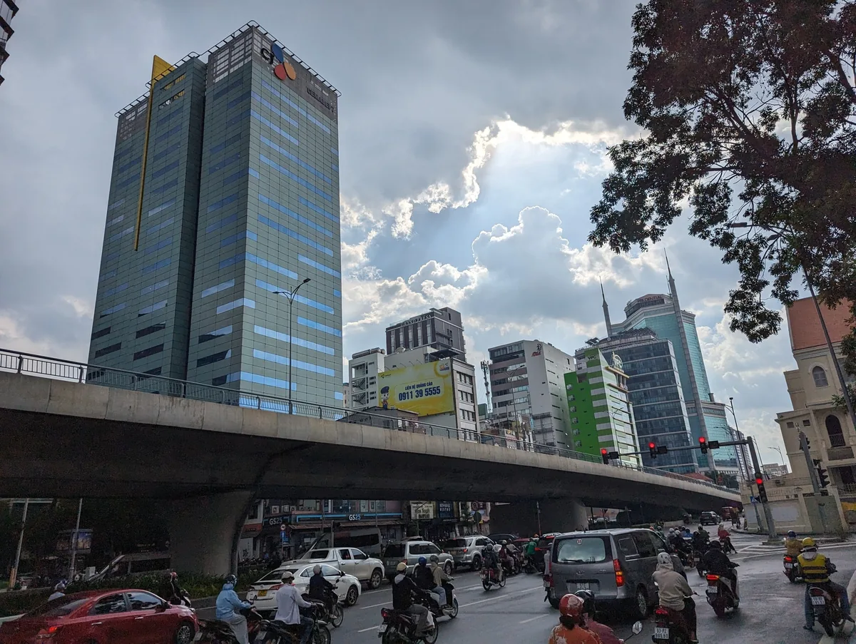 An overpass crosses a busy intersection with high-rise office towers under a cloudy sky in Ho Chi Minh City.