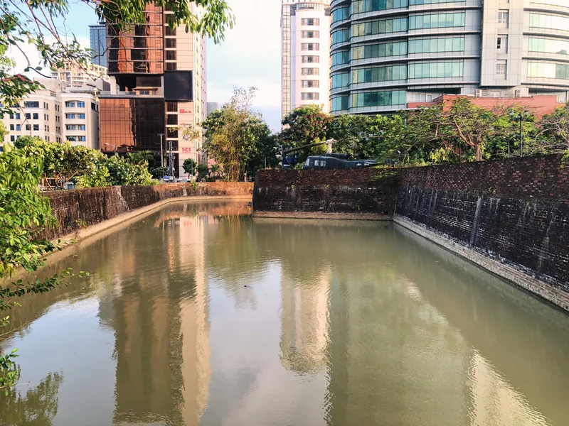 A moat with old brick walls surrounds Dien Hai Citadel, reflecting modern skyscrapers in central Da Nang.