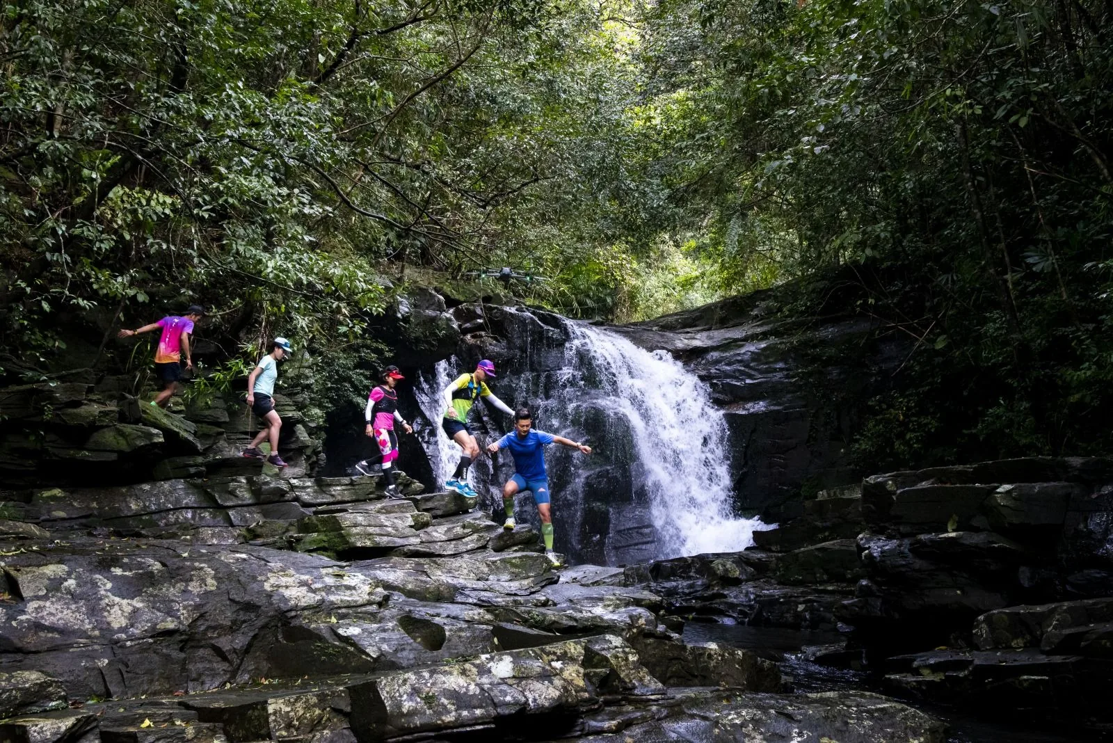 Trail runners scramble over rocky terrain beside a forest waterfall in Bach Ma National Park.